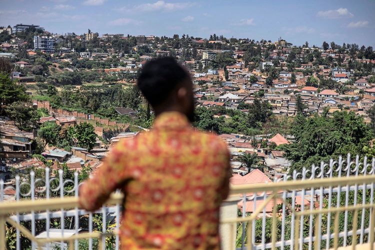 Ein Mann in einem bunten Hemd steht auf einem Balkon mit Blick auf eine hügelige Stadtlandschaft, die dicht mit Häusern und Bäumen bebaut ist. Der Fokus liegt auf der Aussicht; die Person ist unscharf im Vordergrund zu sehen.