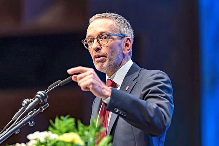 Ein Mann im Anzug und mit Brille spricht gestikulierend an einem Mikrofon. Im Vordergrund sind Blumen sichtbar, der Hintergrund ist unscharf und dunkelblau beleuchtet.