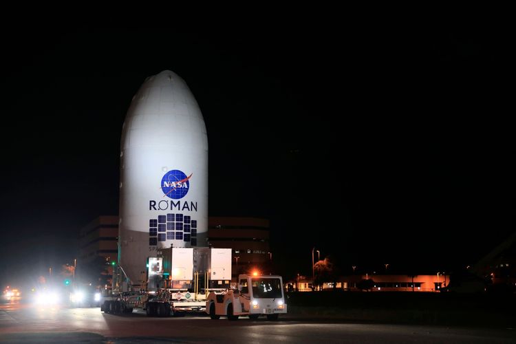 Das Bild zeigt das Nancy Grace Roman Weltraumteleskop der NASA, das in seiner Nutzlastverkleidung eingekapselt ist. Es wird in der Nacht von einem Transportfahrzeug vom Payload Hazardous Servicing Facility zur SpaceX-Halle am Startkomplex 39A im Kennedy Space Center der NASA transportiert. Das Teleskop soll mit einer SpaceX Falcon Heavy Rakete gestartet werden. Im Hintergrund sind beleuchtete Gebäude und Straßenlaternen zu sehen.