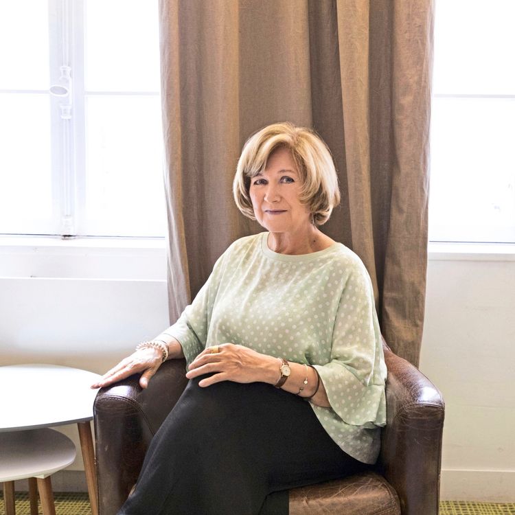 Die Portugiesische Schriftstellerin Lidia Jorge mit blonden halblangen Haaren sitzt in einem braunen Sessel vor einem Fenster mit Vorhängen. Sie trägt eine hellgrüne, gepunktete Bluse und ein dunkles Unterteil.