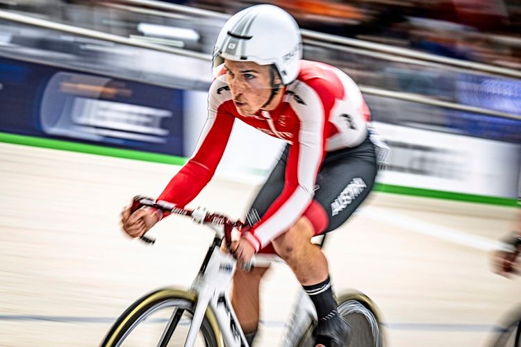 Ein Bahnradfahrer in einem rot-weißen Rennanzug und weißem Helm fährt auf einer Indoor-Radrennbahn. Der Hintergrund ist durch die Bewegung verschwommen, was die Geschwindigkeit betont.