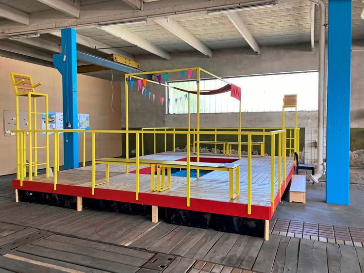 Eine Indoor-Plattform aus Holz mit Geländern in Gelb, umgeben von dekorativen Wimpelketten. Sie steht in einer Halle mit sichtbaren blauen Stützpfeilern und einer hohen Decke. Darunter befindet sich ein kleiner Bereich mit blauen Kugeln, ähnlich einem Bällebad.