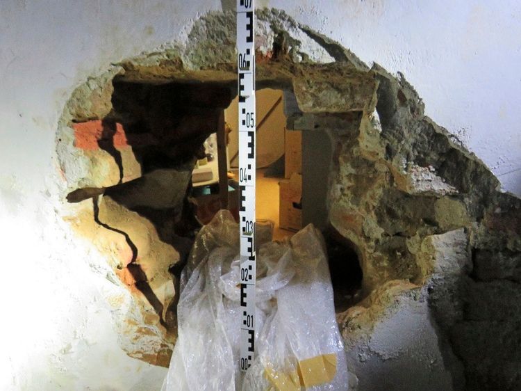 Eine durchbrochene Mauer mit einem großen, unregelmäßigen Loch, das offenbar von Einbrechern geschaffen wurde. Durch das Loch sind Teile eines angrenzenden Raums sichtbar. Vor der Mauer steht eine Maßlatte, und im Vordergrund ist eine Plastikplane erkennbar.