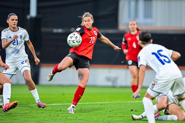 Eine Fußballspielerin im roten Trikot von Österreich kontrolliert den Ball während eines Spiels. Einige Gegenspielerinnen im weißen Trikot des Kosovo beobachten die Szene. Das Spiel findet auf einem Rasenplatz statt.