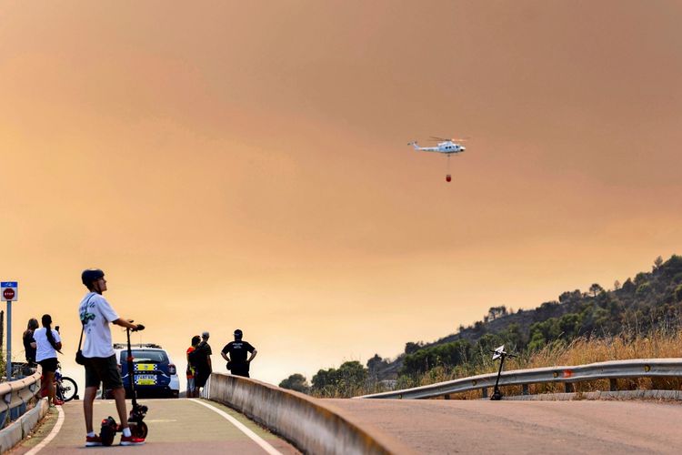 Ein Hubschrauber fliegt mit einem Wasserbehälter über eine von einem Waldbrand betroffene Region in Betxí, Castellón, Valencianische Gemeinschaft, Spanien. Im Vordergrund stehen Menschen auf einer Straße und beobachten die Szene, während der Himmel durch Rauch orange gefärbt ist.