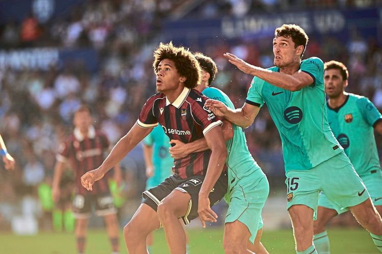 Zwei Fußballspieler kämpfen während eines Spiels um den Ball, einer im rot-schwarz gestreiften Trikot von Levante UD und der andere im türkisfarbenen Trikot des FC Barcelona. Im Hintergrund sind Spieler und Zuschauer unscharf zu erkennen.
