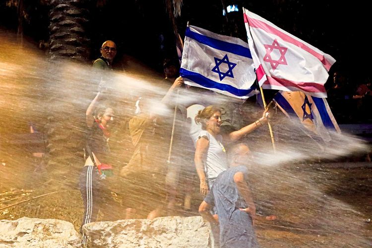 Eine Demonstrantin und ein Demonstrant halten zwei Israel-Flaggen hoch. Sie stehen in einem Wasserstrahl. Die Polizei setzt Wasserwerfer ein.