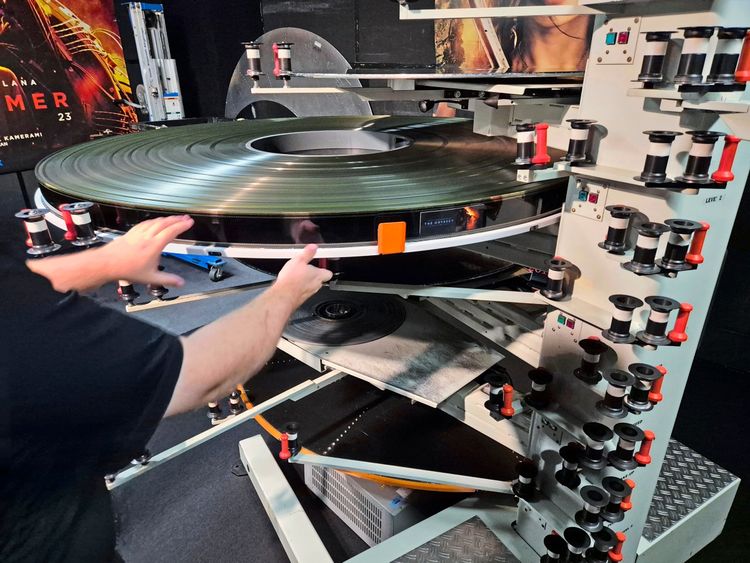 Ein Mensch arbeitet an einer großen Filmrollen-Spulmaschine. Die Maschine hat mehrere Ebenen mit Halterungen für Filmrollen. Im Hintergrund ist ein Filmplakat zu sehen.