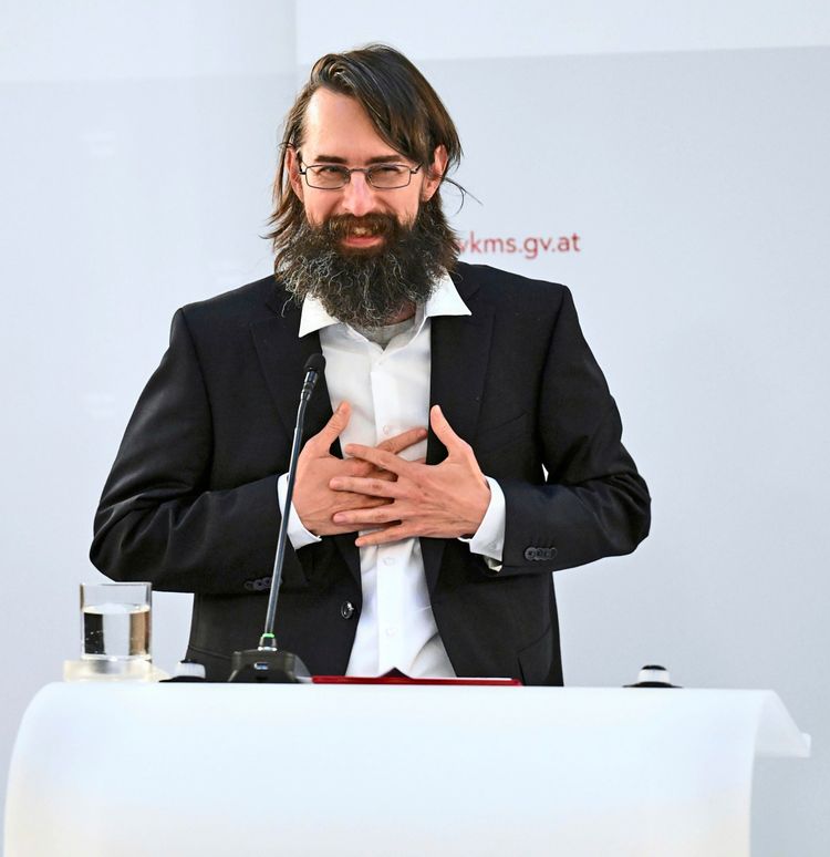 Ein Mann mit Brille, langen Haaren und Vollbart steht an einem Rednerpult. Er trägt einen dunklen Anzug mit weißem Hemd und hält seine Hände auf der Brust. Vor ihm befinden sich ein Mikrofon, ein Glas Wasser und Unterlagen. Im Hintergrund ist eine weiße Wand mit einem Schriftzug sichtbar.