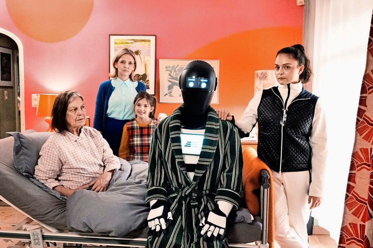 In einem farbenfrohen Raum steht eine Gruppe von Menschen und ein humanoider Roboter zusammen. Eine ältere Frau sitzt in einem Pflegebett, während ein Mädchen, eine Frau und eine weitere Person neben dem Roboter stehen. Der Roboter trägt einen gestreiften Bademantel, und sein Bildschirm zeigt ein neutrales Gesicht mit leuchtenden Augen. Die Stimmung wirkt ruhig und freundschaftlich.