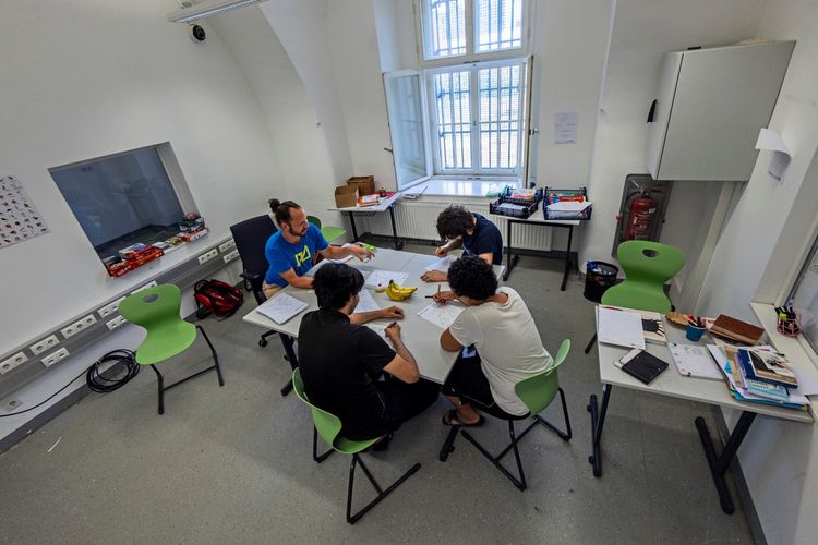 Vier Personen sitzen in einem einfachen, hellen Raum an einem Tisch und arbeiten zusammen an schriftlichen Unterlagen. Im Hintergrund sind ein Fenster, Regale mit Materialien und ein Feuerlöscher zu sehen. Die Szene wirkt wie ein Unterricht oder eine Lernsituation.