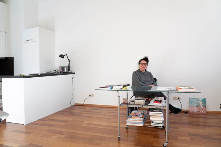 Schweeger sitzt an einem Schreibtisch mit Glasplatte in einem ansonst leeren Raum. Auf dem Tisch liegen Bücher, die Wände sind weiß, der Boden aus rotstichigem Holz.
