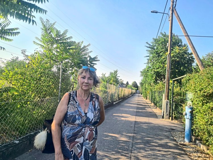 Eine Frau steht auf einem sonnigen Gehweg, der von grünen Pflanzen, einem Maschendrahtzaun und Straßenlaternen gesäumt ist. Sie trägt ein bedrucktes Kleid, Sonnenbrillen auf dem Kopf und eine schwarze Tasche mit einem flauschigen Anhänger. Im Hintergrund erstreckt sich der Weg in die Ferne.