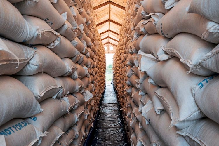 In einem Lagerhaus sind zwei hohe Stapel von weißen Säcken mit Sorghum zu sehen, die bis zur Decke reichen. Die Säcke stammen vom World Food Program und sind für die Verteilung in Maban vorbereitet. Im Hintergrund ist diffuses Tageslicht zu erkennen.