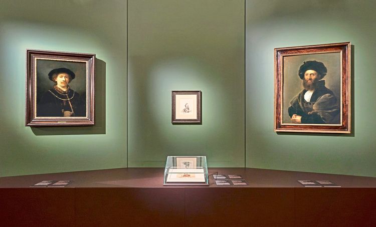 Eine Ausstellungswand mit grünem Hintergrund, auf der zwei gemalte Porträts in goldenen Rahmen hängen. Zwischen den Gemälden befindet sich eine kleinere gerahmte Zeichnung. Auf einer Vitrine darunter liegen weitere Kunstwerke in flacher Präsentation.