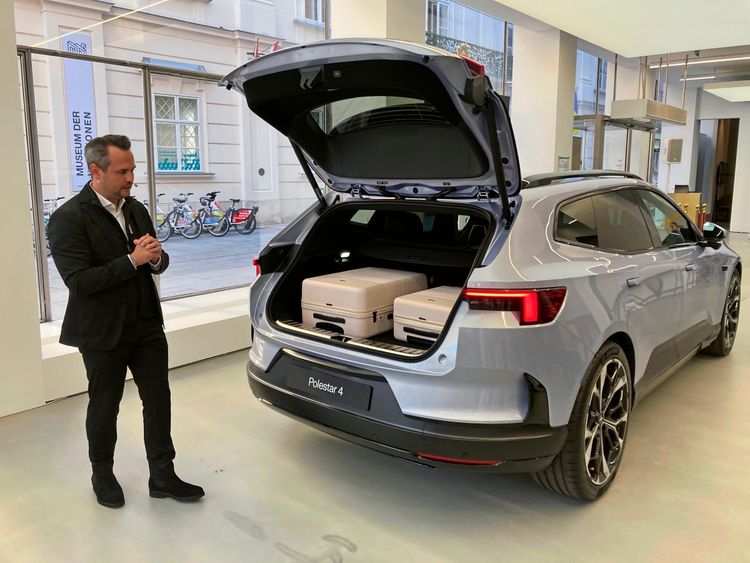Ein silberner Polestar 4 SUV steht mit geöffneter Heckklappe in einem modernen Ausstellungsraum. Zwei Koffer sind im Kofferraum verstaut. Ein Mann in dunkler Kleidung steht links daneben und betrachtet das Fahrzeug. Im Hintergrund sind große Fenster, ein Gebäude und Fahrräder zu sehen.