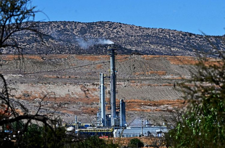 Eine Industrieanlage mit Schornsteinen, aus denen Rauch aufsteigt, ist vor einer trockenen, hügeligen Landschaft in Tiltil, Region Santiago, Chile, zu sehen. Im Vordergrund befinden sich Bäume und Vegetation. Die Aufnahme stammt vom 23. April 2026.