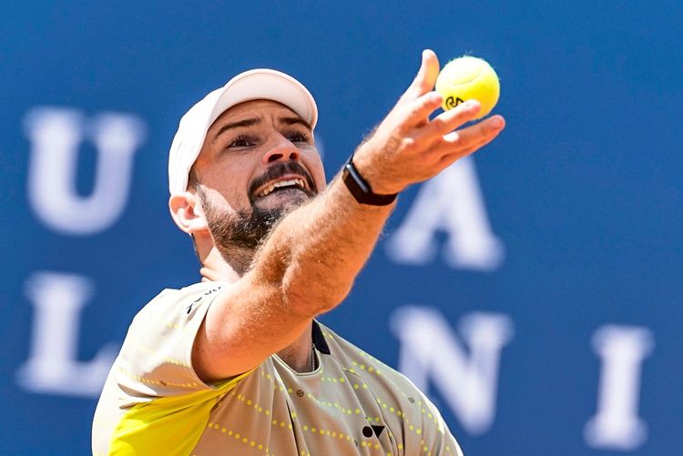 Ein Tennisspieler in Aktion während eines Aufschlags bei einem ATP-Tennisturnier. Der Spieler trägt ein helles Trikot und eine weiße Kappe, im Hintergrund ein blauer Banner.