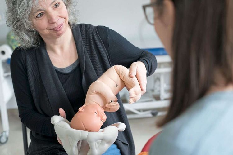 Hebamme hält eine Gebärmutter aus Plastik und eine Puppe in der Hand. Sie erklärt einer jungen, werdenden Mama den Geburtsprozess.
