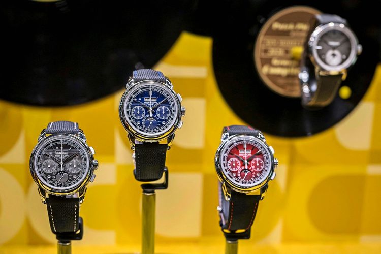 Drei Luxusuhren der Marke Patek Philippe mit unterschiedlichen Zifferblättern in Schwarz, Blau und Rot werden auf Ständern ausgestellt. Im Hintergrund sind weitere Uhren sowie eine gelbe Wand mit geometrischem Muster zu sehen. Die Aufnahme stammt von der Uhrenmesse 