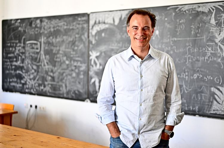 Ein Mann steht in einem Klassenzimmer vor einer großen Tafel, die mit komplexen Formeln und Skizzen beschrieben ist. Er trägt ein helles Hemd und hat die Hände in den Hosentaschen. Sein Gesicht ist unkenntlich gemacht.