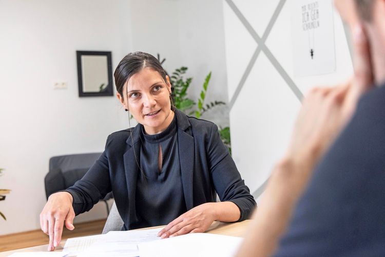 Eine Frau in schwarzer Kleidung sitzt an einem Tisch in einem Büro und zeigt auf Dokumente vor ihr. Im Hintergrund sind Pflanzen, ein Spiegel und eine Wanddekoration zu sehen.