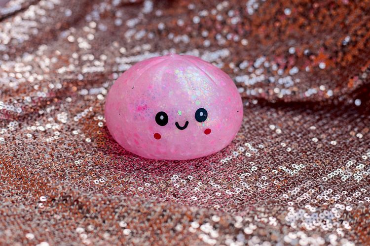 Rosa, glitzerndes Stressabbau-Spielzeug in Form eines Knödels mit einem niedlichen, lächelnden Gesicht, platziert auf einem funkelnden roségoldenen Paillettenhintergrund.