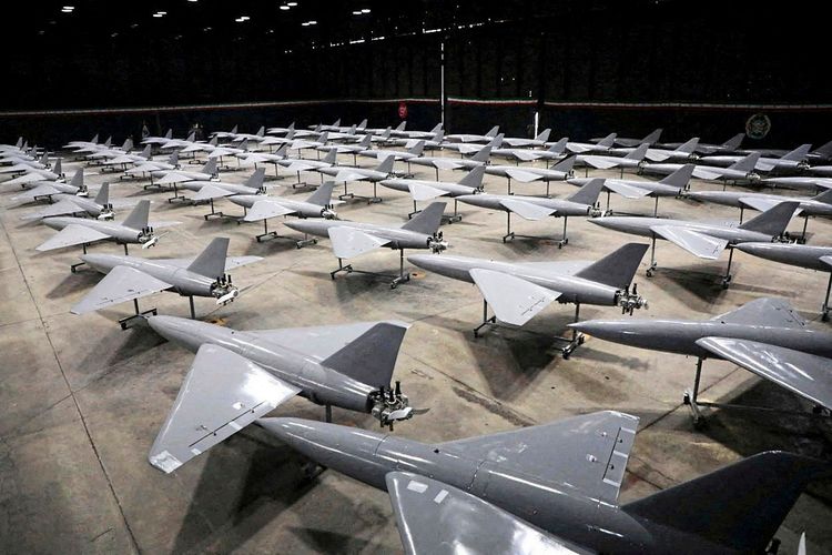 Eine große Anzahl grauer Drohnen ist in mehreren Reihen in einem Hangar aufgestellt. Die Drohnen stehen auf Metallgestellen, und der Raum ist schwach beleuchtet.
