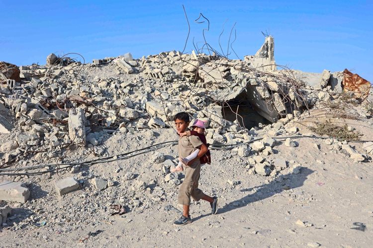 Ein palästinensischer Junge trägt ein kleines Mädchen auf seinem Rücken und geht an den Trümmern zerstörter Gebäude im Flüchtlingslager Jabalia im nördlichen Gazastreifen vorbei. Der Boden ist voller Schutt, und im Hintergrund sind Überreste von Gebäuden und Stahlträgern zu sehen. Der Himmel ist klar und blau.