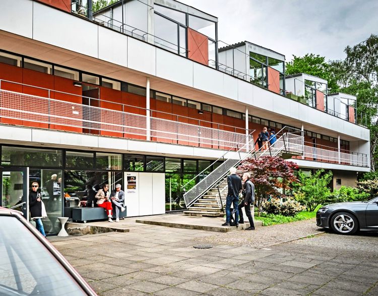 Mehrfamilienhaus im Hansaviertel, bekannt als Eternithaus von Paul Baumgarten, mit moderner Architektur, roten Fassadenelementen und Balkonen. Menschen sind vor dem Gebäude zu sehen, einige sitzen auf einer Bank, andere stehen in Gruppen. Im Vordergrund parken Autos. Fotografiert während des Gallery Weekend 2025 in Berlin.
