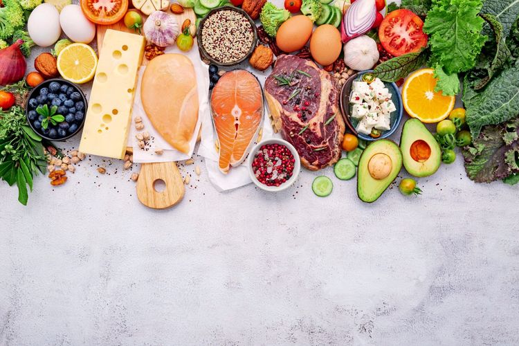 Wichtige Ingedienzien eine Keto-Diät:  Lachs, Hähnchenfilet, Steak, Eier, Käse, Avocado, Blattgemüse, Tomaten, Gurken, Heidelbeeren, Nüsse, Samen, Feta-Käse, Kräuter, Zitrone und Orange, dekorativ auf einem weißen Hintergrund arrangiert.