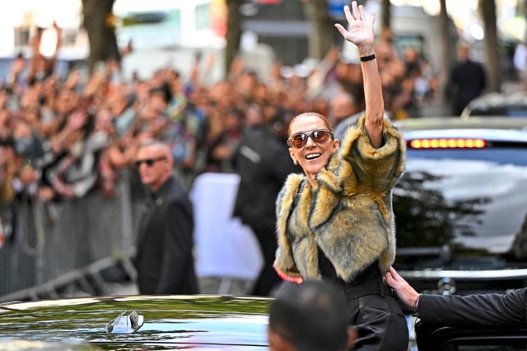 Eine Frau mit Sonnenbrille und einem Fellmantel winkt lächelnd einer großen Menschenmenge zu, während sie neben einem Auto steht. Im Hintergrund sind Fans hinter Absperrungen zu sehen.