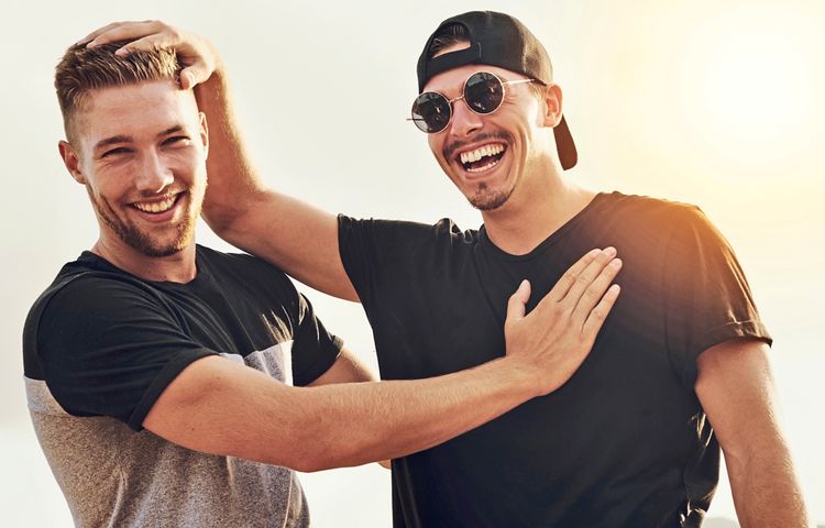 Zwei lachende junge Männer stehen draußen, einer trägt eine Sonnenbrille und eine umgedrehte Kappe, während sie sich freundschaftlich umarmen und Spaß haben.