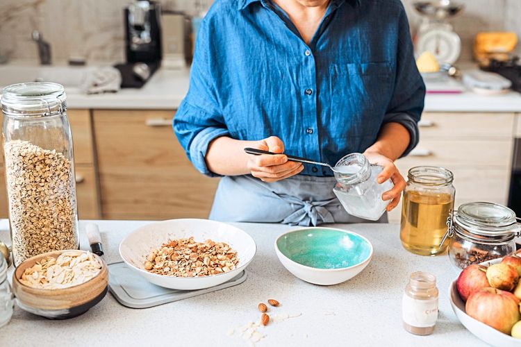 Frau mit blauem Hemd in der Küche, die Granola vorbereitet und gerade einen Löffel Kokosöl in eine Schüssel gibt