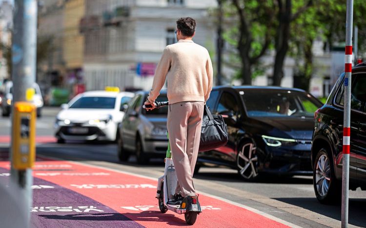 Ein Mann fährt mit einem E-Scooter auf einem rot markierten Radweg in einer Stadt. Autos fahren daneben auf der Straße. Straßenszene bei Tageslicht mit moderner urbaner Kulisse.