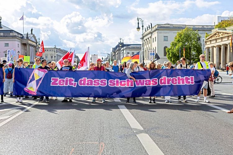 Eine Gruppe von Demonstrierenden hält ein großes blaues Banner mit der Aufschrift „Zeit, dass sich was dreht!“ in pinker Schrift, auf dem auch eine stilisierte Sanduhr abgebildet ist. Im Hintergrund sind weitere Demonstrierende mit Plakaten und Flaggen zu sehen sowie Gebäude in Berlin.