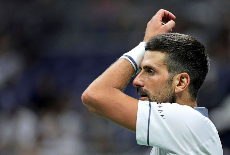 Ein Tennisspieler im weißen Trikot mit Schweißband am Handgelenk streicht sich die Haare aus dem Gesicht bei einem Match. Der Hintergrund ist unscharf, die Szene findet in einem Stadion statt.