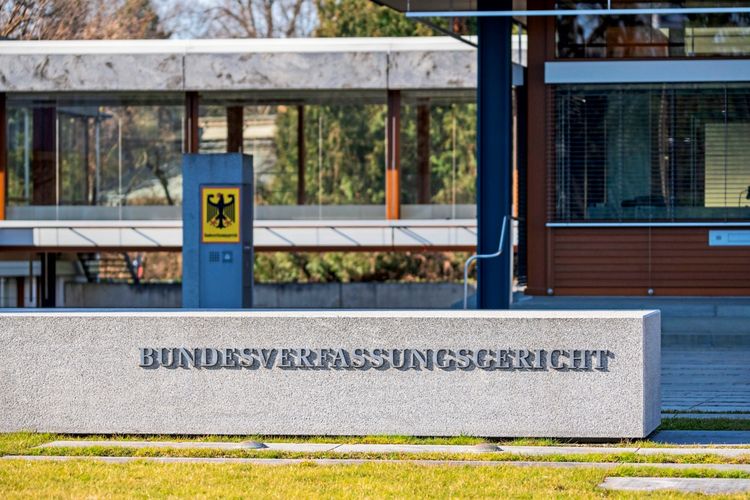 Ein massiver Steinblock mit der Aufschrift „BUNDESVERFASSUNGSGERICHT“ steht vor dem Gebäude des Gerichts in Karlsruhe. Im Hintergrund ist das moderne Gebäude mit Glasfassade zu sehen, daneben das Wappen der Bundesrepublik Deutschland mit dem Bundesadler.