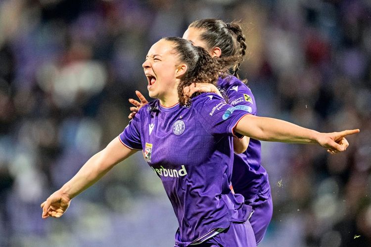 Eine Fußballspielerin im Trikot von FK Austria Wien jubelt enthusiastisch mit ausgestreckten Armen nach einem Tor. Eine Mitspielerin umarmt sie von hinten. Im Hintergrund unscharfe Zuschauertribüne.