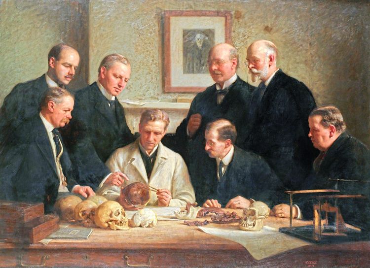 Gemälde von Wissenschaftern in schwarzen Anzügen, die um einen Kollegen mit weißem Labormantel stehen, der verschiedene Schädel vor sich auf dem Tisch liegen hat. Zur dargestellten 