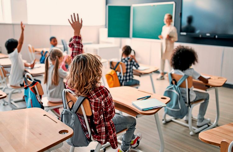 Mehrere Grundschulkinder sitzen in einem Klassenzimmer an Holzschreibtischen. Einige Kinder haben die Hand gehoben, um sich zu melden. Im Hintergrund steht die Lehrerin vor einer Tafel. Schultaschen hängen an den Stühlen, und die Atmosphäre ist lebhaft.