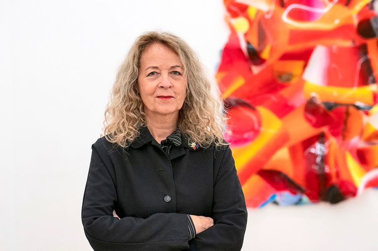 Eine Frau mit schulterlangen, lockigen, blonden Haaren steht mit verschränkten Armen vor einer bunten, modernen Kunstinstallation. Sie trägt eine schwarze Jacke, und der Hintergrund ist weiß.