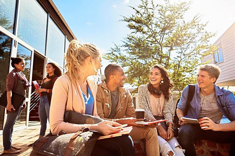 Eine bunt gemischte Gruppe junger Menschen sitzt lachend vor einem Gebäude auf einem Uni-Campus