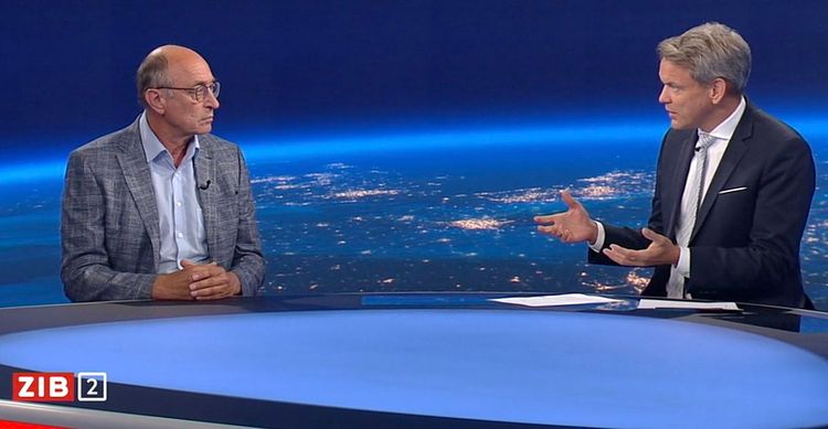 Mann mit Glatze, Brille und grauem Sakko sitzt links im Bild am blauen Studiopult der ZIB 2 einem gestikulierenden Mann mit grauem Haar und dunklem Sakko vor dem nachtblauen Studiohintergrund der 