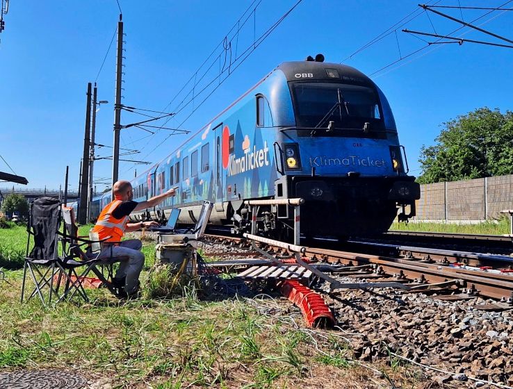 Ein Zug der ÖBB mit der Aufschrift „KlimaTicket“ fährt auf Gleisen entlang. Im Vordergrund sitzt eine Person in einer orangenen Warnweste auf einem Klappstuhl neben den Gleisen. Der Himmel ist klar und blau, im Hintergrund sind Strommasten und eine Lärmschutzwand sichtbar.