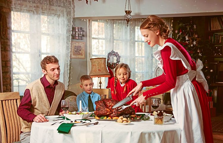 Familie mit zwei Kindern sitzt am Tisch, die Mutter schneidet den Truthahn auf, im Hintergrund ein Weihnachtsbaum