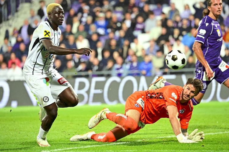 Ein Fußballspiel in der Generali-Arena in Wien: Ein Spieler von Sturm Graz (links) und der Torwart von Austria Wien (in orange) sind in Aktion, während der Ball in der Luft ist. Im Hintergrund sind Zuschauer und ein weiterer Spieler zu sehen.