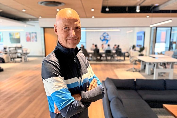 Ein Mann steht in einem modernen Büro mit Holzboden. Er trägt eine sportliche Jacke mit blauen und weißen Streifen und hat die Arme verschränkt. Im Hintergrund sind ein Sofa, ein Couchtisch und ein Besprechungsraum mit Menschen sichtbar. Auf dem Boden steht eine weiße Tasse.