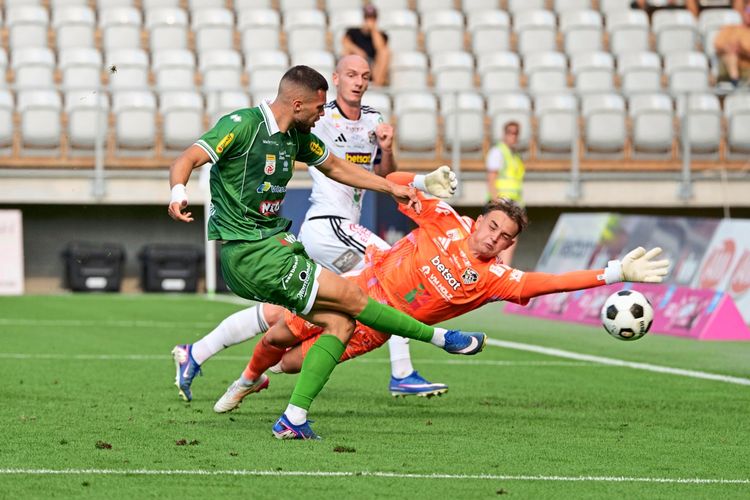 Ein Fußballspieler in grün (SC Austria Lustenau) versucht, den Ball am Torwart in orange (RZ Pellets WAC) vorbeizuspielen. Szene aus einem Bundesligaspiel, mit leeren Tribünen im Hintergrund.
