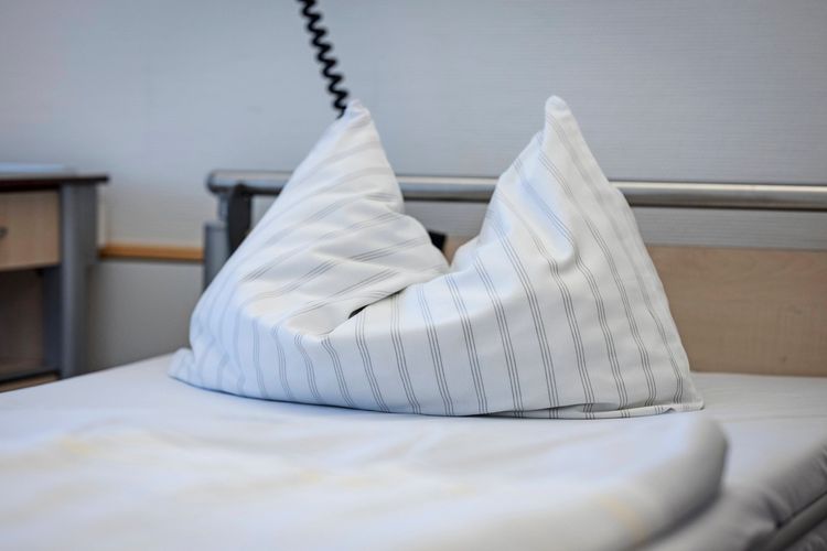 Ein weißes Kissen mit dezentem Streifenmuster liegt auf einem Krankenbett in einem Krankenhauszimmer. Im Hintergrund sind ein Nachttisch und ein Teil des Bettrahmens sichtbar.